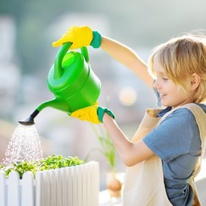 Cute boy is watering self-grown microgreens plants in a box on the balcony on sunny summer day